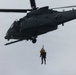 A dark gray military helicopter, identifiable as a U.S. Air Force HH-60 Pave Hawk, is hovering against an overcast sky. A person is being hoisted on a cable beneath the aircraft, wearing a yellow flotation device and helmet, indicating a search and rescue or combat rescue training operation. The scene depicts a hoist/rescue extraction exercise or live rescue mission.