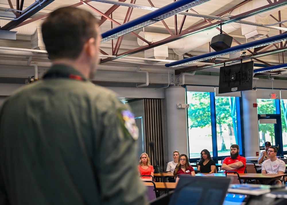 A person wearing a military flight suit with a patch on the back is presenting or speaking to a small group of civilians seated at desks in what appears to be a modern classroom or briefing room. The audience of approximately six people is attentively listening to the presenter. The setting features industrial-style exposed ceiling infrastructure with blue beams and a ceiling-mounted display screen.