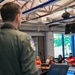 A person wearing a military flight suit with a patch on the back is presenting or speaking to a small group of civilians seated at desks in what appears to be a modern classroom or briefing room. The audience of approximately six people is attentively listening to the presenter. The setting features industrial-style exposed ceiling infrastructure with blue beams and a ceiling-mounted display screen.