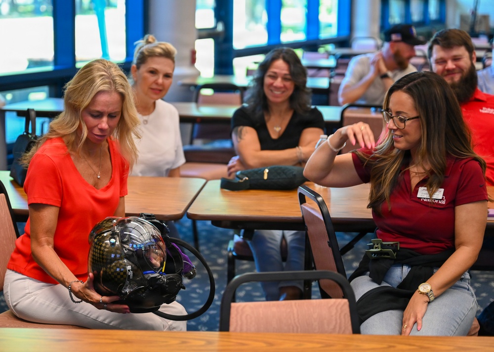 A group of people are gathered in what appears to be a conference or training room, examining a specialized helmet or breathing apparatus. A woman in a red top is holding and inspecting a complex black helmet with attached hoses and a visor, while a woman in a maroon polo shirt with a name badge looks on. Several other attendees are seated at tables in the background, observing the demonstration.