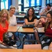 A group of people are gathered in what appears to be a conference or training room, examining a specialized helmet or breathing apparatus. A woman in a red top is holding and inspecting a complex black helmet with attached hoses and a visor, while a woman in a maroon polo shirt with a name badge looks on. Several other attendees are seated at tables in the background, observing the demonstration.