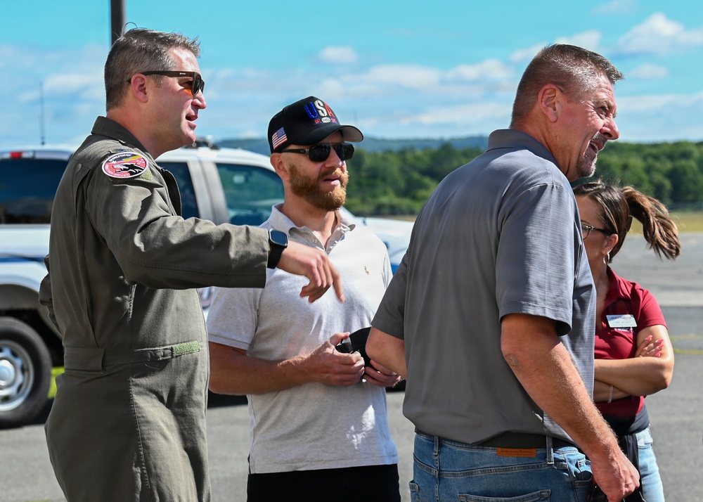 A military pilot in an olive green flight suit is conversing with a small group of civilians on what appears to be an airport tarmac or airfield. The pilot, wearing sunglasses and a squadron patch on his arm, appears to be gesturing and explaining something to the group. The setting suggests a military open house, air show, or VIP visit event.