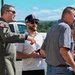 A military pilot in an olive green flight suit is conversing with a small group of civilians on what appears to be an airport tarmac or airfield. The pilot, wearing sunglasses and a squadron patch on his arm, appears to be gesturing and explaining something to the group. The setting suggests a military open house, air show, or VIP visit event.