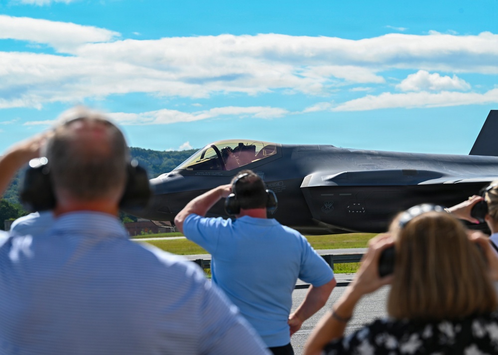 A group of spectators wearing hearing protection observe a military stealth fighter jet, consistent with an F-35 Lightning II, taxiing on a runway. The scene appears to be at an airshow or military base open house event on a sunny day. At least one spectator is photographing the aircraft with a camera.