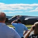 A group of spectators wearing hearing protection observe a military stealth fighter jet, consistent with an F-35 Lightning II, taxiing on a runway. The scene appears to be at an airshow or military base open house event on a sunny day. At least one spectator is photographing the aircraft with a camera.