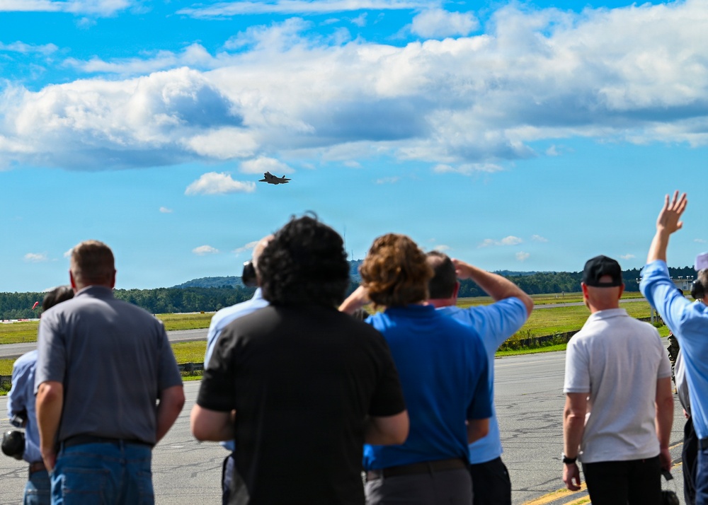 A group of spectators stands on an airport tarmac watching a military jet aircraft in flight overhead. The observers are facing away from the camera, with several shielding their eyes or raising hands as they track the aircraft against a partly cloudy blue sky. The setting appears to be an airfield observation event or air show.