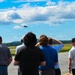 A group of spectators stands on an airport tarmac watching a military jet aircraft in flight overhead. The observers are facing away from the camera, with several shielding their eyes or raising hands as they track the aircraft against a partly cloudy blue sky. The setting appears to be an airfield observation event or air show.