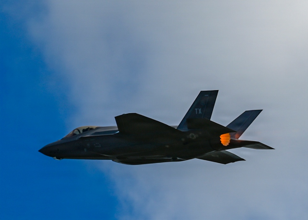 A dark-colored F-35 stealth fighter jet is captured in flight against a partly cloudy blue sky. The aircraft displays 'TX' tail code markings, indicating it belongs to a Texas-based unit, likely Luke Air Force Base or NAS JRB Fort Worth. The engine afterburner is visibly ignited, producing an orange flame at the rear of the aircraft.