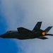 A dark-colored F-35 stealth fighter jet is captured in flight against a partly cloudy blue sky. The aircraft displays 'TX' tail code markings, indicating it belongs to a Texas-based unit, likely Luke Air Force Base or NAS JRB Fort Worth. The engine afterburner is visibly ignited, producing an orange flame at the rear of the aircraft.