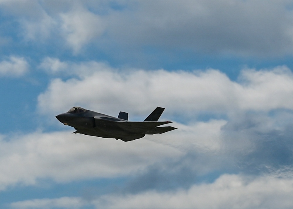 A single stealth fighter jet, consistent with an F-35 Lightning II, is captured in flight against a partly cloudy blue sky. The aircraft appears to be in level flight or a slight climb, with visible vapor trails streaming from its wingtips. The image was taken from a ground-level perspective looking upward.