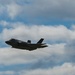 A single stealth fighter jet, consistent with an F-35 Lightning II, is captured in flight against a partly cloudy blue sky. The aircraft appears to be in level flight or a slight climb, with visible vapor trails streaming from its wingtips. The image was taken from a ground-level perspective looking upward.