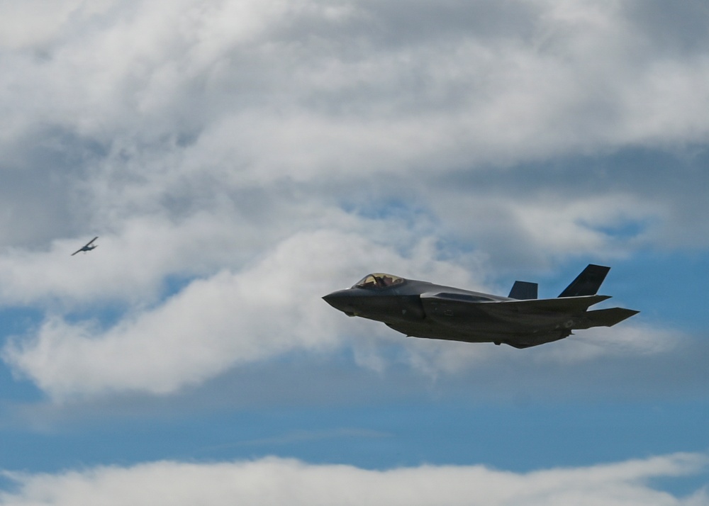 The image shows a modern stealth fighter jet, consistent with an F-35 Lightning II, flying in the foreground against a partly cloudy sky. A second, smaller aircraft is visible in the distant background to the left. The scene appears to be an airshow or military flight demonstration.