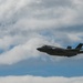 The image shows a modern stealth fighter jet, consistent with an F-35 Lightning II, flying in the foreground against a partly cloudy sky. A second, smaller aircraft is visible in the distant background to the left. The scene appears to be an airshow or military flight demonstration.