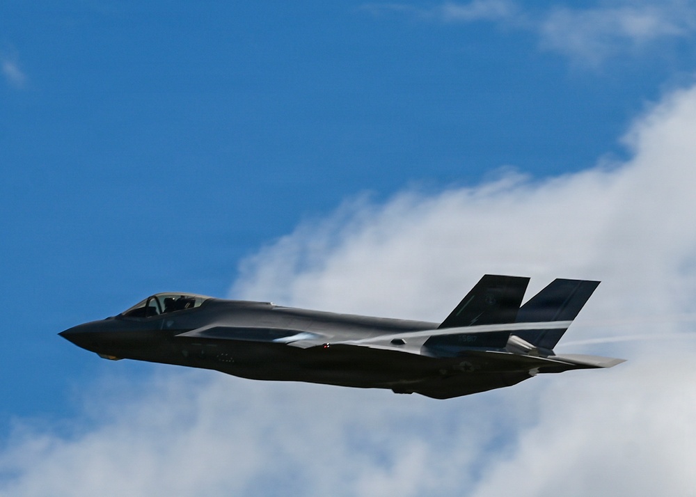 A single stealth fighter jet, consistent with an F-35 Lightning II, is captured in flight against a partly cloudy blue sky. The aircraft is photographed from a slightly low angle, showing its distinctive stealthy profile with twin canted tail fins and a single-seat cockpit. The image appears to have been taken at an airshow or during a demonstration flight.