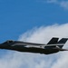 A single stealth fighter jet, consistent with an F-35 Lightning II, is captured in flight against a partly cloudy blue sky. The aircraft is photographed from a slightly low angle, showing its distinctive stealthy profile with twin canted tail fins and a single-seat cockpit. The image appears to have been taken at an airshow or during a demonstration flight.