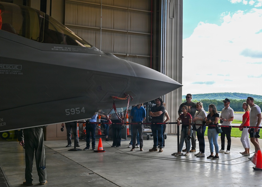 A group of civilians is viewing a military fighter jet, identified by the number '5954' on its fuselage, inside a large aircraft hangar. The aircraft appears to be an F-35 Lightning II based on its distinctive stealth geometry and canopy design. The visitors are standing behind a rope barrier while what appears to be a military personnel member in flight suit stands nearby, suggesting an organized tour or public outreach event.