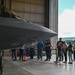 A group of civilians is viewing a military fighter jet, identified by the number '5954' on its fuselage, inside a large aircraft hangar. The aircraft appears to be an F-35 Lightning II based on its distinctive stealth geometry and canopy design. The visitors are standing behind a rope barrier while what appears to be a military personnel member in flight suit stands nearby, suggesting an organized tour or public outreach event.