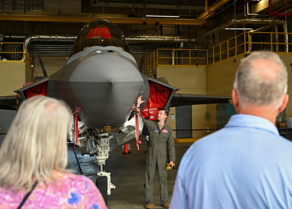 A military pilot in an olive green flight suit is briefing two civilian visitors inside an aircraft hangar, gesturing toward a parked F-35 Lightning II stealth fighter jet. The aircraft is fitted with red 'Remove Before Flight' safety covers and tags on various components. The setting appears to be an official tour or familiarization event at a military air base.