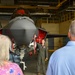 A military pilot in an olive green flight suit is briefing two civilian visitors inside an aircraft hangar, gesturing toward a parked F-35 Lightning II stealth fighter jet. The aircraft is fitted with red 'Remove Before Flight' safety covers and tags on various components. The setting appears to be an official tour or familiarization event at a military air base.