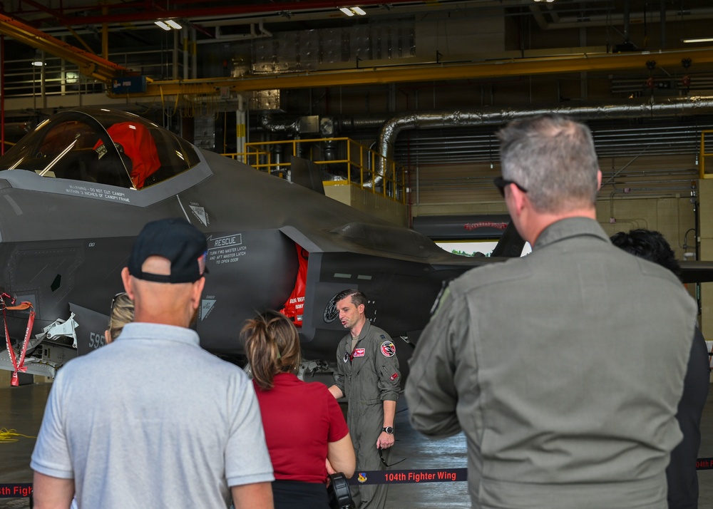 A military pilot in a flight suit is briefing a small group of civilians and at least one other uniformed personnel inside an aircraft hangar. The briefing is taking place in front of an F-35 Lightning II stealth fighter jet, with a banner visible reading '104th Fighter Wing'. This appears to be an official tour or familiarization event at a military installation.