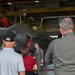 A military pilot in a flight suit is briefing a small group of civilians and at least one other uniformed personnel inside an aircraft hangar. The briefing is taking place in front of an F-35 Lightning II stealth fighter jet, with a banner visible reading '104th Fighter Wing'. This appears to be an official tour or familiarization event at a military installation.