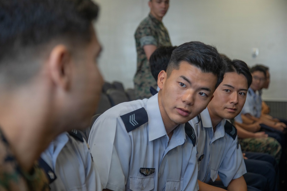 The image shows several young military personnel seated in what appears to be a briefing or meeting room. The individuals in the foreground are wearing light blue/white uniforms with rank insignia on the shoulders, consistent with an air force uniform style. In the background, at least one person is wearing a camouflage uniform, suggesting a multi-branch or joint military gathering.