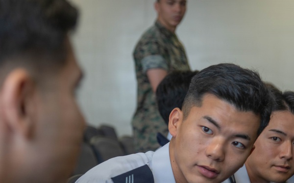 The image shows several young military personnel seated in what appears to be a briefing or meeting room. The individuals in the foreground are wearing light blue/white uniforms with rank insignia on the shoulders, consistent with an air force uniform style. In the background, at least one person is wearing a camouflage uniform, suggesting a multi-branch or joint military gathering.