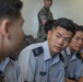 The image shows several young military personnel seated in what appears to be a briefing or meeting room. The individuals in the foreground are wearing light blue/white uniforms with rank insignia on the shoulders, consistent with an air force uniform style. In the background, at least one person is wearing a camouflage uniform, suggesting a multi-branch or joint military gathering.