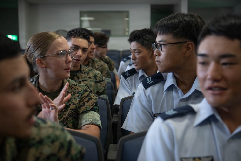 The image shows a group of military personnel in an indoor setting, appearing to be engaged in a discussion or exchange. A female U.S. Marine in digital camouflage uniform is speaking and gesturing with her hands toward individuals wearing light blue/grey uniforms with rank insignia, suggesting a bilateral or multinational military interaction. The setting appears to be a briefing room or classroom environment.