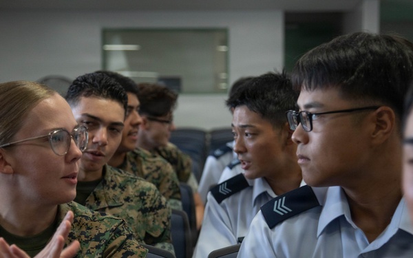 The image shows a group of military personnel in an indoor setting, appearing to be engaged in a discussion or exchange. A female U.S. Marine in digital camouflage uniform is speaking and gesturing with her hands toward individuals wearing light blue/grey uniforms with rank insignia, suggesting a bilateral or multinational military interaction. The setting appears to be a briefing room or classroom environment.