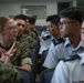 The image shows a group of military personnel in an indoor setting, appearing to be engaged in a discussion or exchange. A female U.S. Marine in digital camouflage uniform is speaking and gesturing with her hands toward individuals wearing light blue/grey uniforms with rank insignia, suggesting a bilateral or multinational military interaction. The setting appears to be a briefing room or classroom environment.