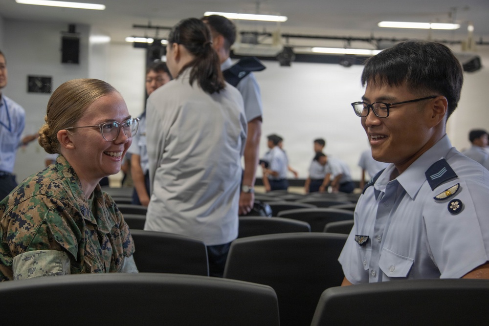 Two military personnel in different uniforms are engaged in a friendly conversation in what appears to be a briefing room or auditorium. The person on the left wears US Marine Corps digital camouflage (MARPAT), while the person on the right wears a light blue uniform consistent with an Asian air force service. Several other uniformed individuals are visible in the background.