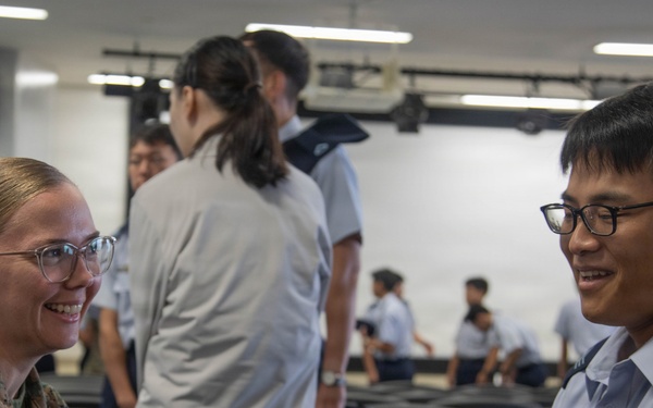 Two military personnel in different uniforms are engaged in a friendly conversation in what appears to be a briefing room or auditorium. The person on the left wears US Marine Corps digital camouflage (MARPAT), while the person on the right wears a light blue uniform consistent with an Asian air force service. Several other uniformed individuals are visible in the background.