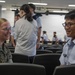 Two military personnel in different uniforms are engaged in a friendly conversation in what appears to be a briefing room or auditorium. The person on the left wears US Marine Corps digital camouflage (MARPAT), while the person on the right wears a light blue uniform consistent with an Asian air force service. Several other uniformed individuals are visible in the background.