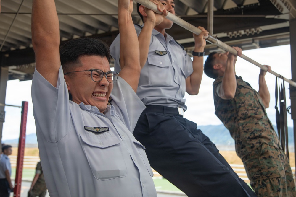 Multiple military personnel are performing pull-ups on a horizontal bar under a covered outdoor structure. Two individuals are wearing light blue uniforms with wing insignia badges, while a third person on the right is wearing woodland camouflage (MARPAT-style) uniform. The foreground subject is visibly straining with an intense facial expression while wearing glasses.