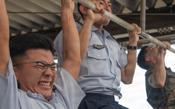 Multiple military personnel are performing pull-ups on a horizontal bar under a covered outdoor structure. Two individuals are wearing light blue uniforms with wing insignia badges, while a third person on the right is wearing woodland camouflage (MARPAT-style) uniform. The foreground subject is visibly straining with an intense facial expression while wearing glasses.