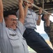 Multiple military personnel are performing pull-ups on a horizontal bar under a covered outdoor structure. Two individuals are wearing light blue uniforms with wing insignia badges, while a third person on the right is wearing woodland camouflage (MARPAT-style) uniform. The foreground subject is visibly straining with an intense facial expression while wearing glasses.