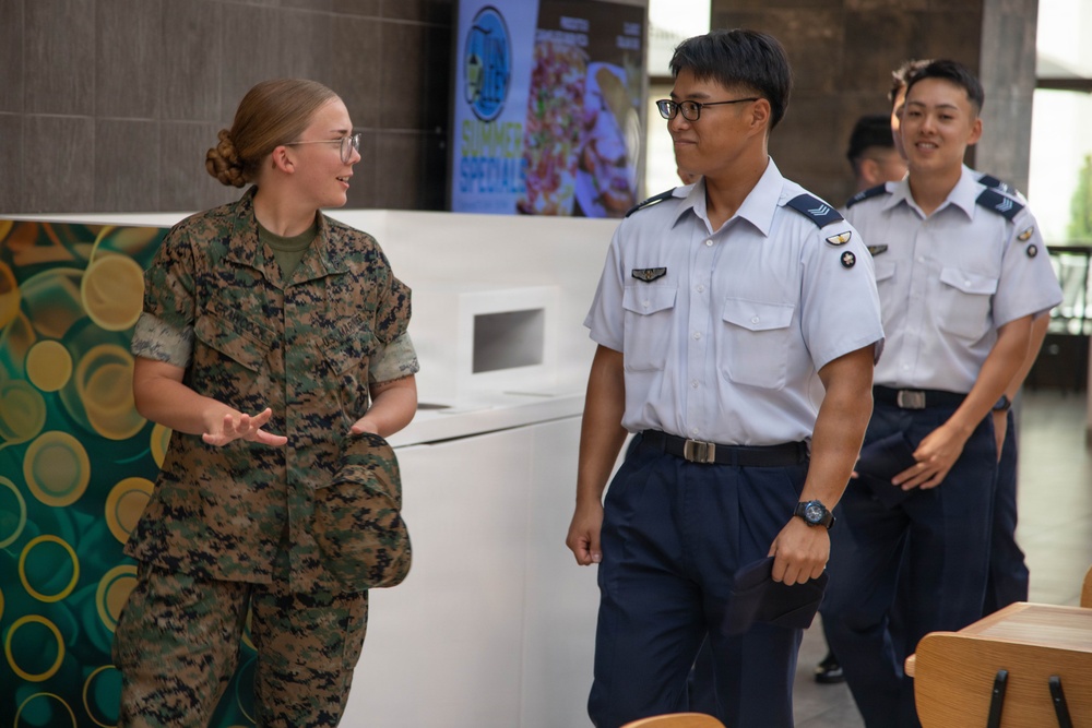 A U.S. Marine in woodland MARPAT camouflage uniform is engaged in conversation with military personnel wearing light blue and navy dress uniforms, likely from an Asian air force. The interaction appears friendly and informal, possibly during a joint military exchange or liaison event. The setting appears to be an indoor facility, possibly a dining facility or common area.