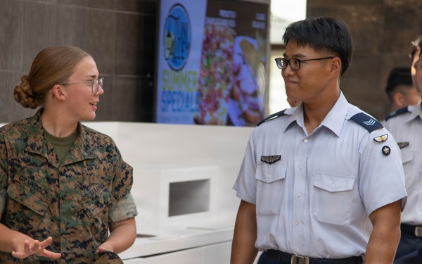 A U.S. Marine in woodland MARPAT camouflage uniform is engaged in conversation with military personnel wearing light blue and navy dress uniforms, likely from an Asian air force. The interaction appears friendly and informal, possibly during a joint military exchange or liaison event. The setting appears to be an indoor facility, possibly a dining facility or common area.