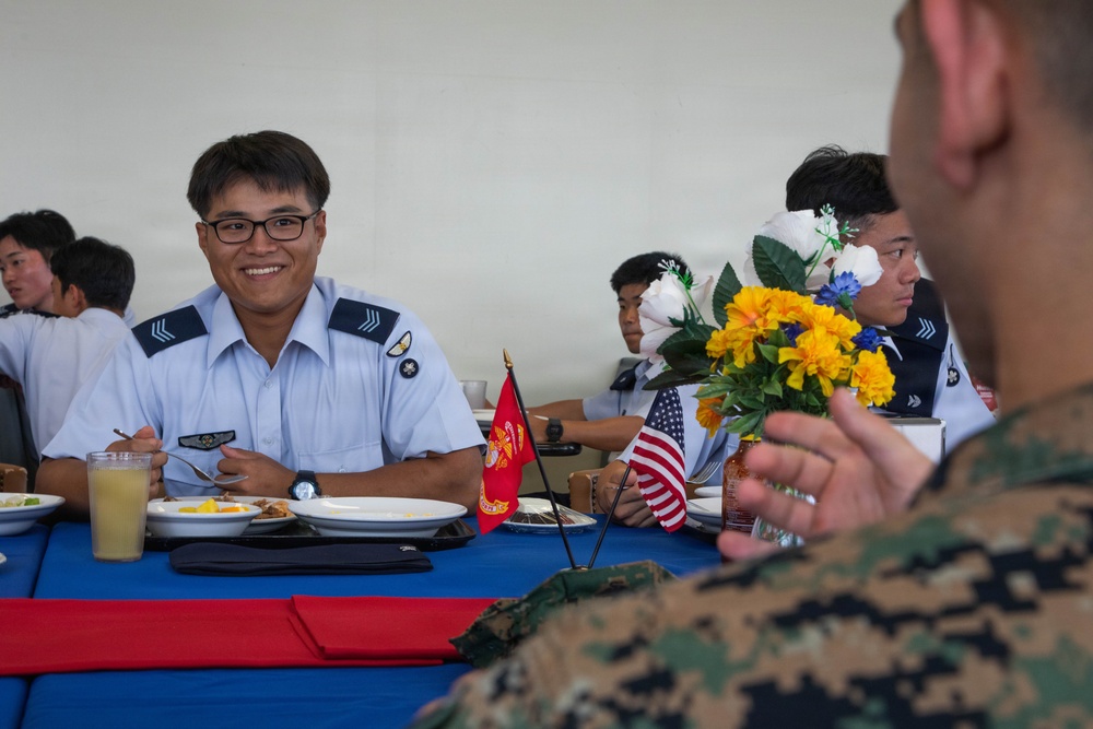 Military personnel from different services are sharing a meal together at a table decorated with American flags, a Marine Corps flag, and flower arrangements. The setting appears to be a formal military dining event or exchange program, with personnel wearing light blue uniforms (consistent with an Asian air force) seated across from a U.S. Marine in MARPAT camouflage. The atmosphere is friendly and conversational, with one servicemember smiling broadly at the camera.