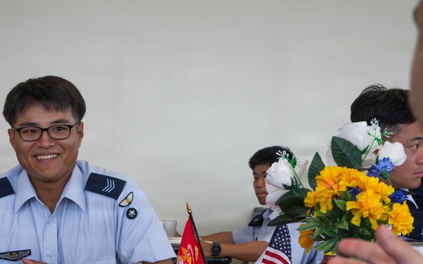 Military personnel from different services are sharing a meal together at a table decorated with American flags, a Marine Corps flag, and flower arrangements. The setting appears to be a formal military dining event or exchange program, with personnel wearing light blue uniforms (consistent with an Asian air force) seated across from a U.S. Marine in MARPAT camouflage. The atmosphere is friendly and conversational, with one servicemember smiling broadly at the camera.