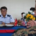 Military personnel from different services are sharing a meal together at a table decorated with American flags, a Marine Corps flag, and flower arrangements. The setting appears to be a formal military dining event or exchange program, with personnel wearing light blue uniforms (consistent with an Asian air force) seated across from a U.S. Marine in MARPAT camouflage. The atmosphere is friendly and conversational, with one servicemember smiling broadly at the camera.