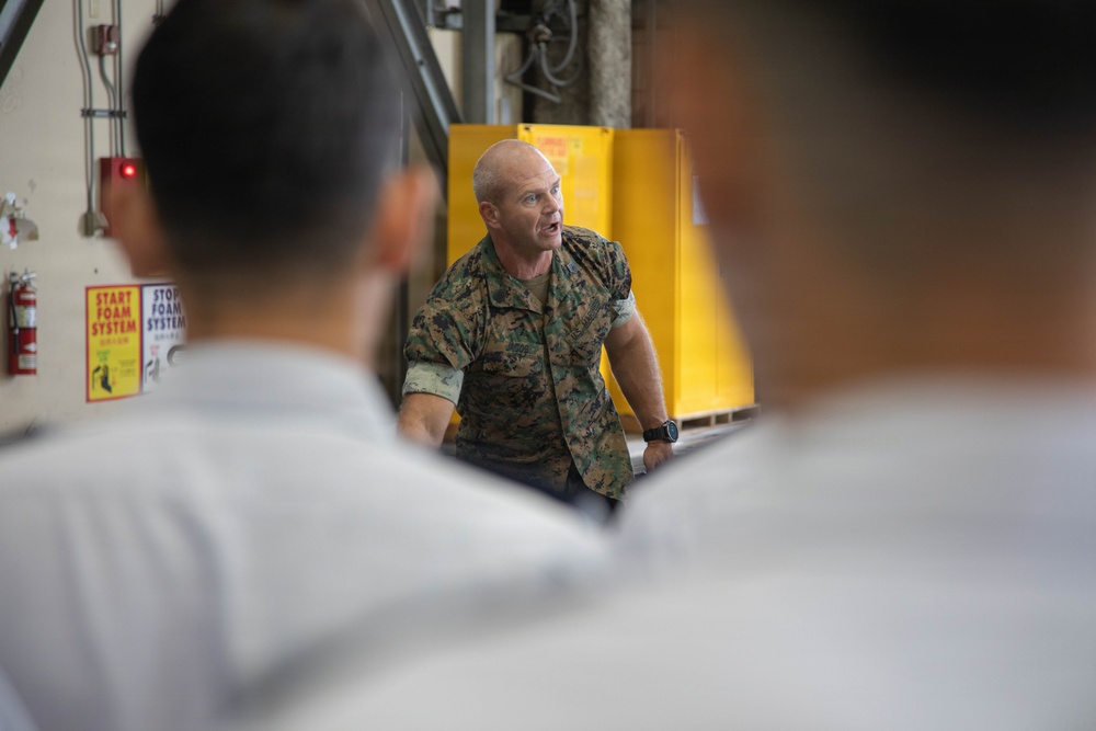 A military personnel member in camouflage uniform appears to be briefing or instructing a group of individuals in what appears to be an industrial or hangar facility. The speaker is addressing an audience whose backs are visible in the foreground, suggesting a formal instruction or orientation session. The setting includes safety signage and industrial equipment in the background.