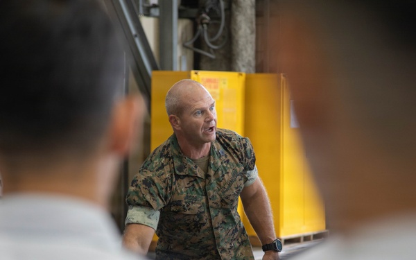 A military personnel member in camouflage uniform appears to be briefing or instructing a group of individuals in what appears to be an industrial or hangar facility. The speaker is addressing an audience whose backs are visible in the foreground, suggesting a formal instruction or orientation session. The setting includes safety signage and industrial equipment in the background.