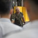 A military personnel member in camouflage uniform appears to be briefing or instructing a group of individuals in what appears to be an industrial or hangar facility. The speaker is addressing an audience whose backs are visible in the foreground, suggesting a formal instruction or orientation session. The setting includes safety signage and industrial equipment in the background.