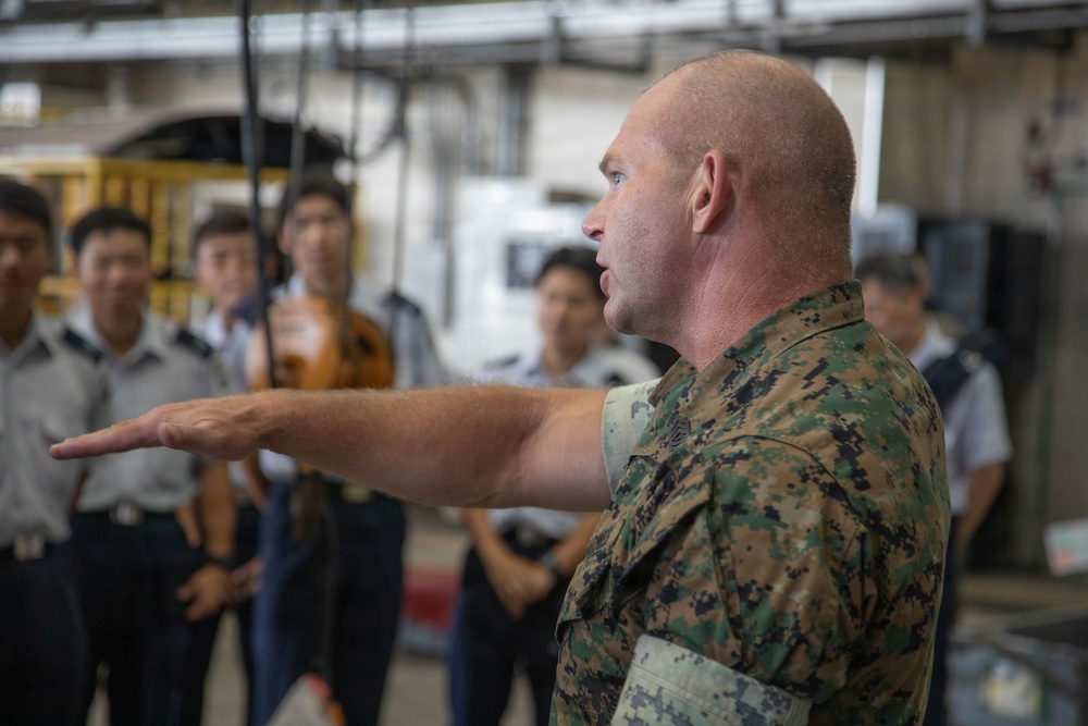 A U.S. Marine in woodland MARPAT camouflage uniform is gesturing with an outstretched arm while apparently briefing or instructing a group of individuals in light gray uniforms, likely foreign military or government personnel. The setting appears to be an industrial or hangar-type facility. The scene suggests an official military exchange, tour, or training event.