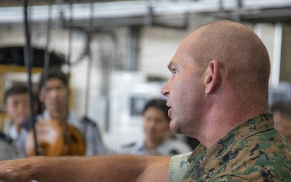 A U.S. Marine in woodland MARPAT camouflage uniform is gesturing with an outstretched arm while apparently briefing or instructing a group of individuals in light gray uniforms, likely foreign military or government personnel. The setting appears to be an industrial or hangar-type facility. The scene suggests an official military exchange, tour, or training event.