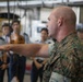 A U.S. Marine in woodland MARPAT camouflage uniform is gesturing with an outstretched arm while apparently briefing or instructing a group of individuals in light gray uniforms, likely foreign military or government personnel. The setting appears to be an industrial or hangar-type facility. The scene suggests an official military exchange, tour, or training event.