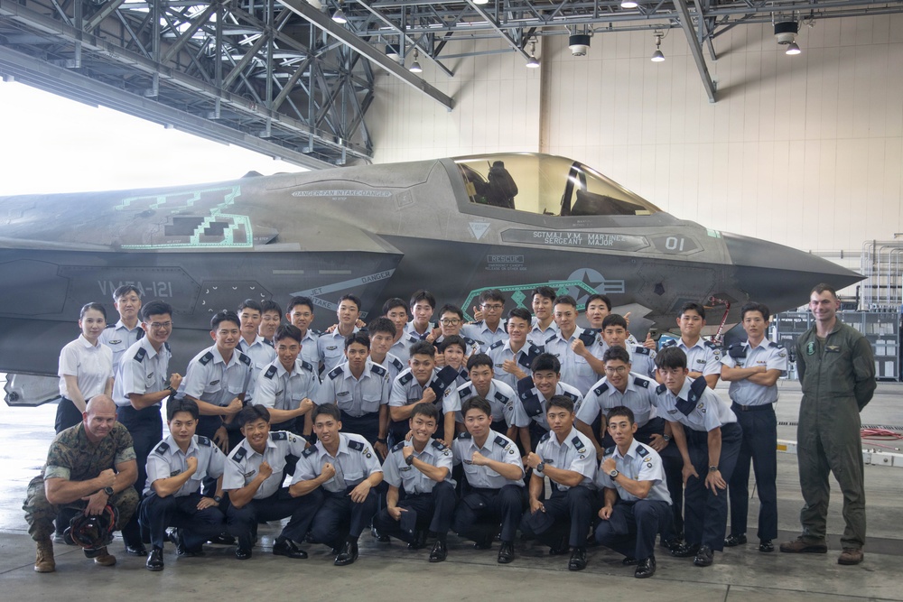 A large group of what appears to be Korean military cadets or students in light blue/grey uniforms pose for a group photo in front of an F-35B fighter jet inside a hangar. The aircraft bears the markings 'VMFA-121', identifying it as belonging to Marine Fighter Attack Squadron 121, a U.S. Marine Corps unit. Two U.S. military personnel — one in camouflage utilities and one in a flight suit — are also present in the photo.