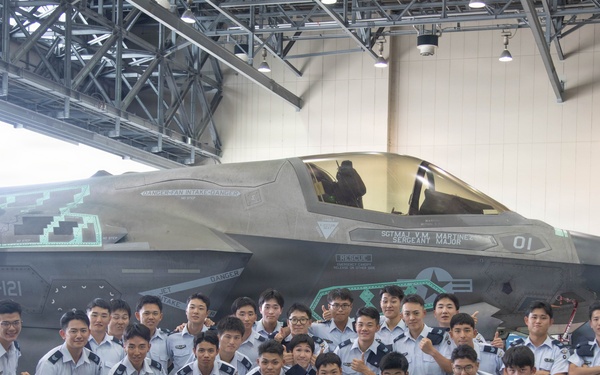 A large group of what appears to be Korean military cadets or students in light blue/grey uniforms pose for a group photo in front of an F-35B fighter jet inside a hangar. The aircraft bears the markings 'VMFA-121', identifying it as belonging to Marine Fighter Attack Squadron 121, a U.S. Marine Corps unit. Two U.S. military personnel — one in camouflage utilities and one in a flight suit — are also present in the photo.